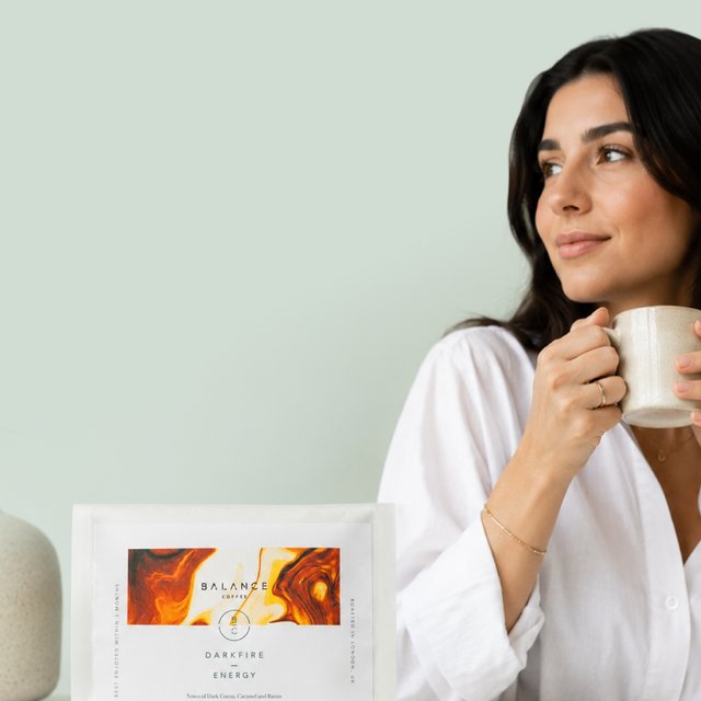 A woman sips coffee from a ceramic mug beside a bag of Balance Coffee Darkfire