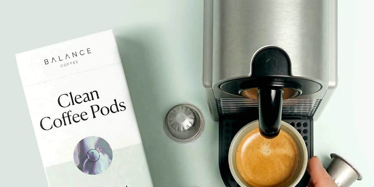A hand placing a cup under a Nespresso machine next to a box of Clean Coffee Pods