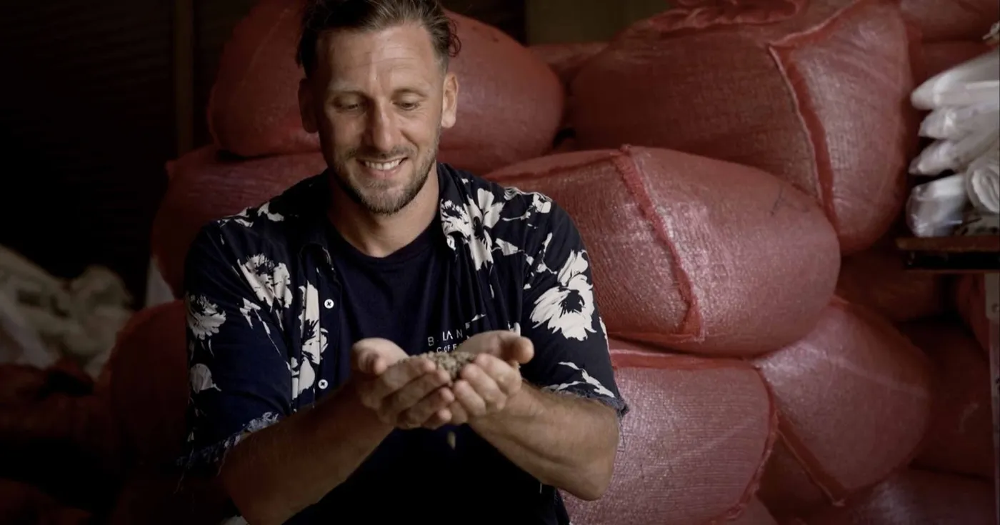 Green coffee beans held in cupped hands in front of stacked coffee sacks