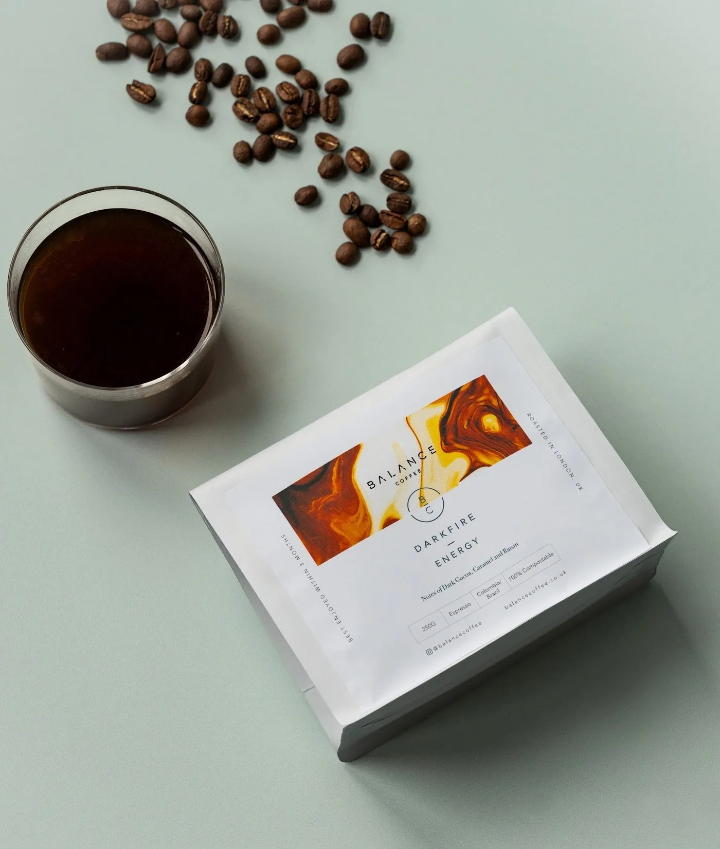 A bag of Balance Darkfire Energy coffee beside a glass of black coffee and scattered beans on a mint background